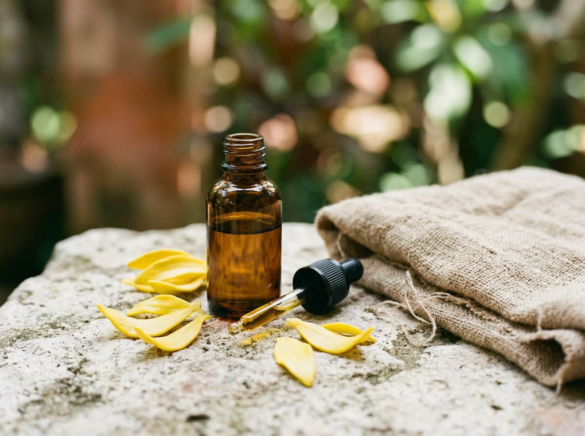 amber dropper bottle of cananga essential oil with petals on linen and stone